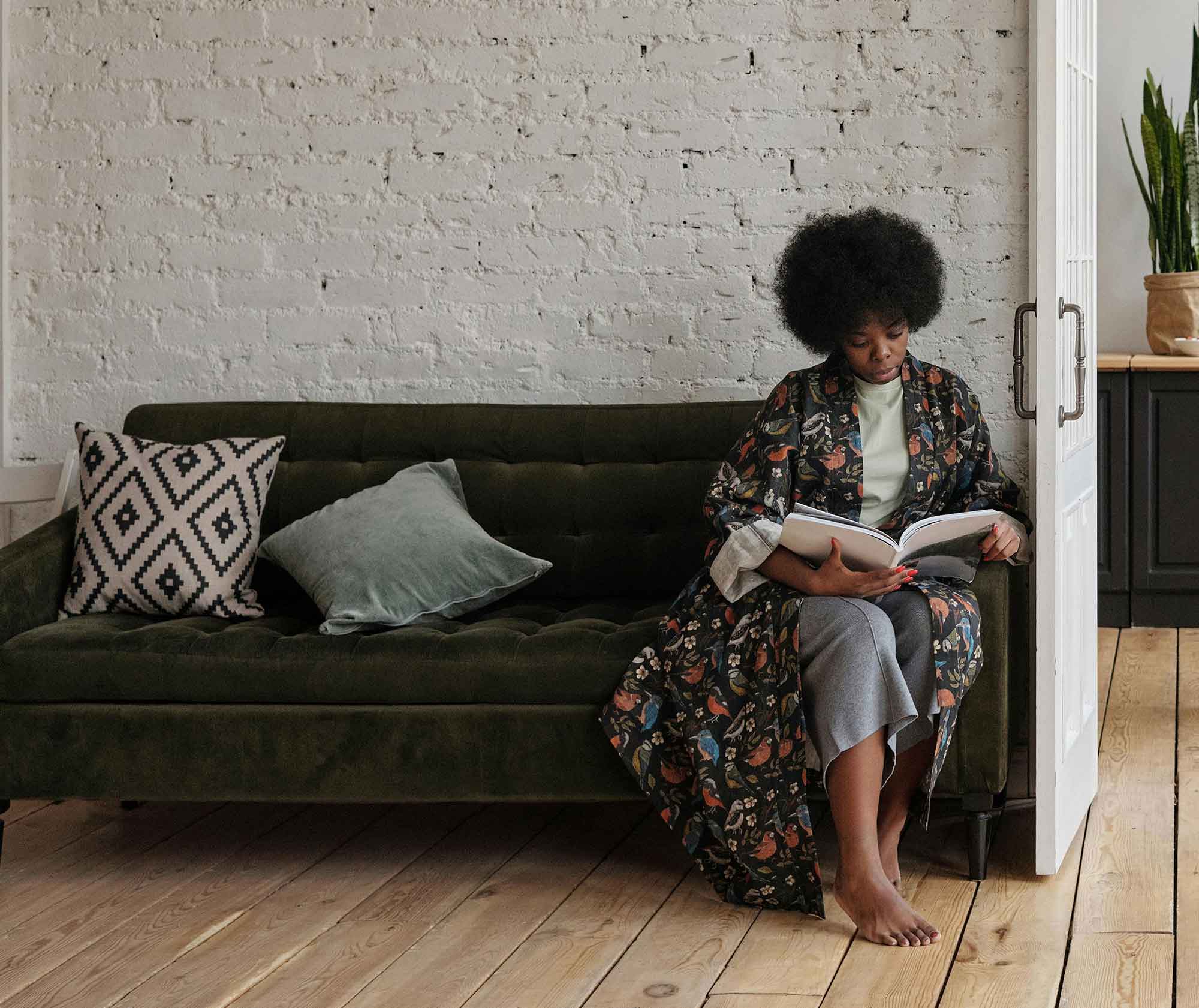 A person is sitting barefoot on a green velvet sofa, deeply engrossed in reading a book, in a minimalist room with a white brick wall.