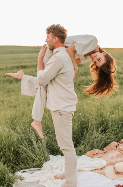 A person is giving a piggyback ride to another person in a sunny, grassy field, near a picnic setup with a blanket and food. Both are smiling.