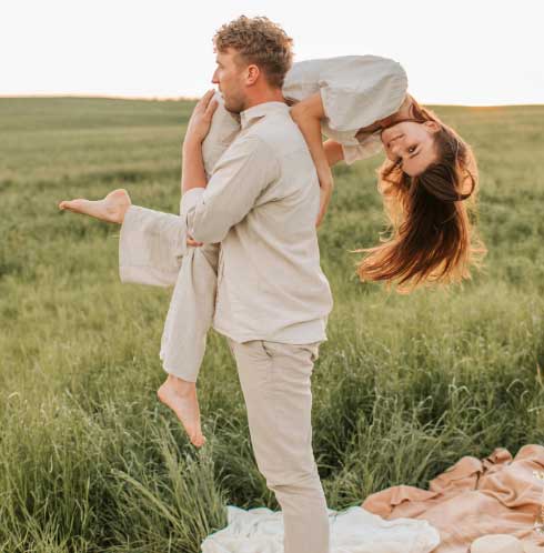 A person carries another upside-down on their back in a grassy field during golden hour, suggesting playfulness and intimacy in a serene outdoor setting.