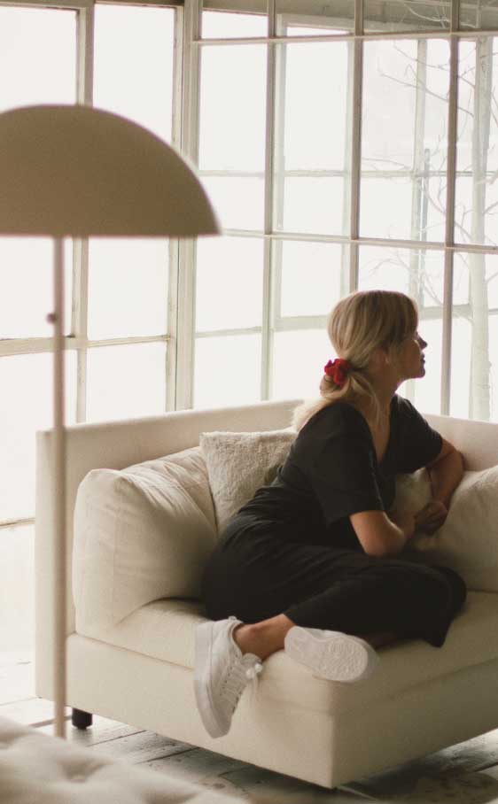 A person is seated on a white sofa, gazing out of large windows, with a red flower in their hair, in a softly lit room with a floor lamp.