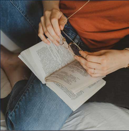 A person sits holding eyeglasses and a paperback book, with text visible. They are wearing a red shirt and denim jeans, casually relaxed.