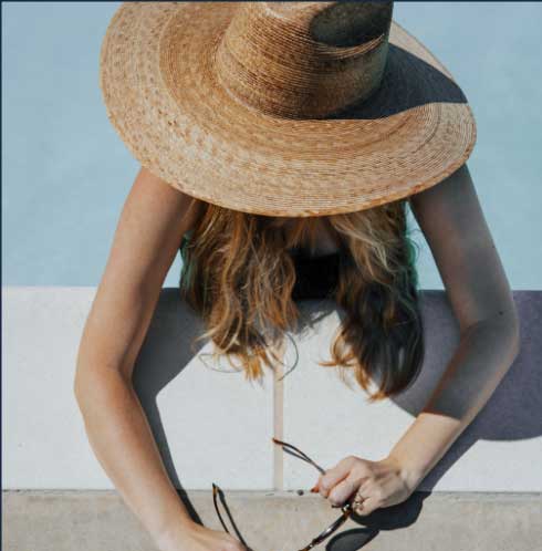 A person wearing a straw hat and holding sunglasses is sitting with their head tilted downwards, obscuring their face, against a pale blue background.