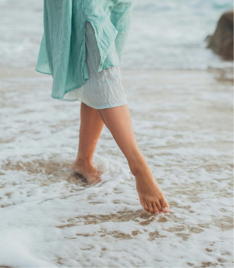 A person is walking along a beach, with a wave washing over their bare feet. They wear a light blue garment, suggesting a serene mood.