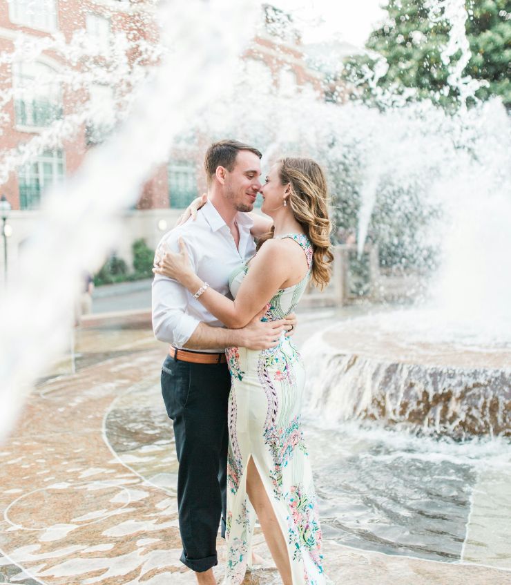 Two people embracing in front of a fountain. The person on the left is wearing a casual shirt and pants, the other a floral dress.