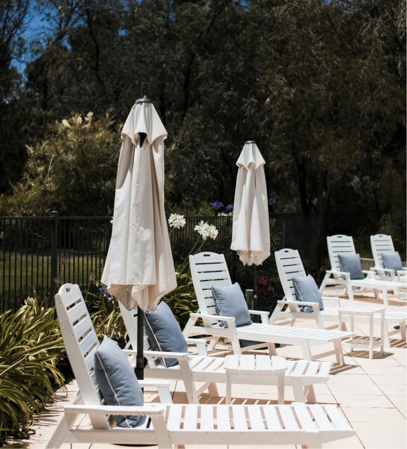 A tranquil outdoor setting with white sun loungers and closed umbrellas arranged neatly on a patio, surrounded by greenery and a fence under a clear sky.