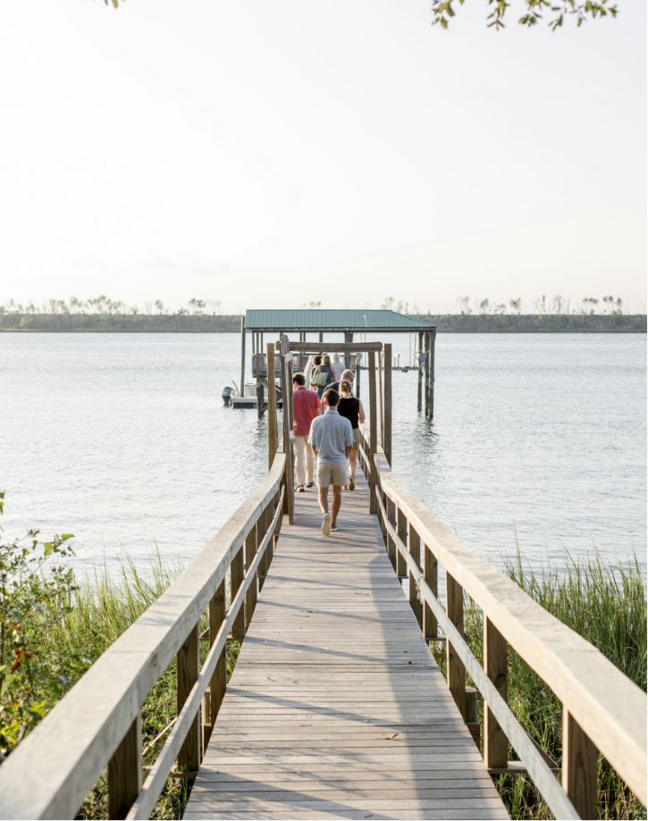 Three people are walking down a wooden pier towards a covered dock on a tranquil body of water, with lush greenery on the left and clear skies above.