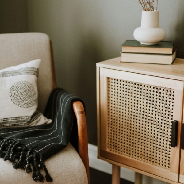 A cozy corner with a beige armchair that has a black throw blanket and an accent pillow. A wooden sideboard with books and a vase rests beside it.