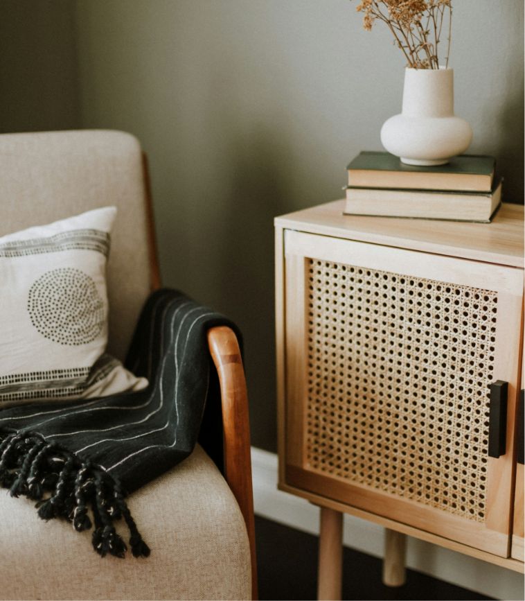 A cozy corner with a beige armchair that has a black throw blanket and an accent pillow. A wooden sideboard with books and a vase rests beside it.