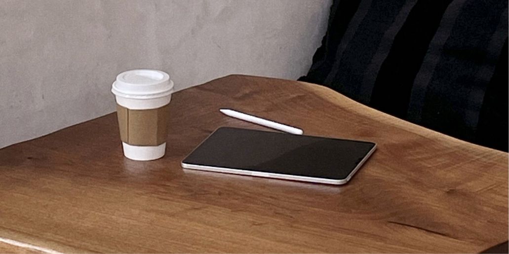 A disposable coffee cup, digital tablet, and stylus lie on a wooden table. There's a hint of a jacket in the background. It suggests a work or study setting.