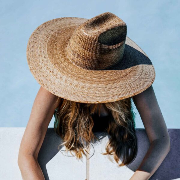 A person is wearing a wide-brimmed straw hat, obscuring their face, with brown wavy hair visible, leaning on a light-colored pool edge.