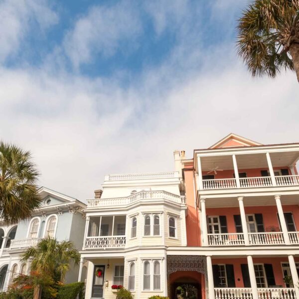 Elegant, colorful houses with multiple stories and balconies line a sunny street, juxtaposed against a blue sky with light clouds, accented by a palm tree.