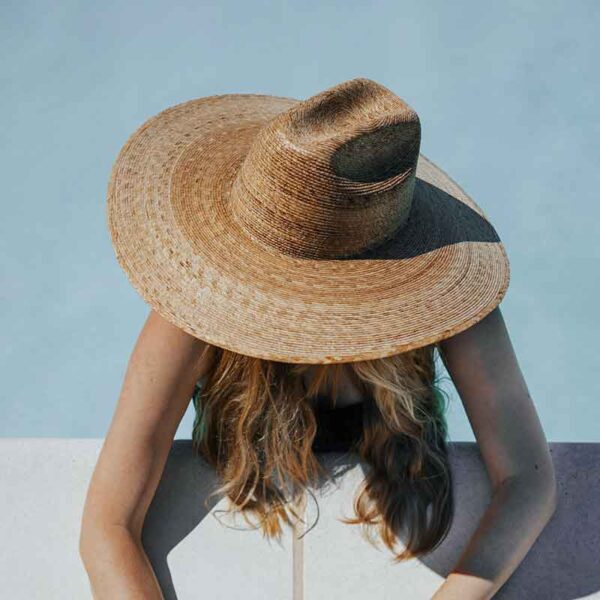 A person is wearing a wide-brimmed straw hat, obscuring their face, positioned above a pool edge with wavy blonde hair draping down.