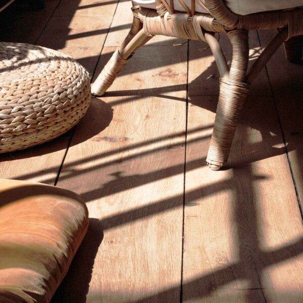 The image shows a warm and cozy interior with sunlight casting shadows, highlighting a woven chair leg, a straw ottoman, and a wooden floor.