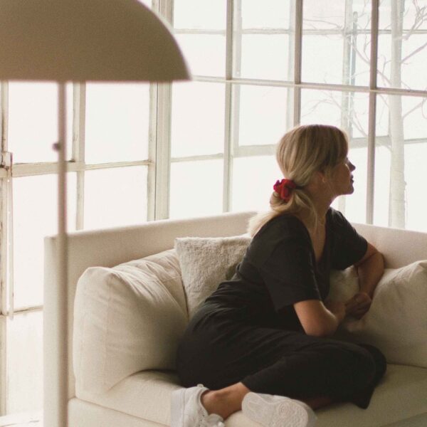 A person with blonde hair wearing a black outfit sits on a white sofa, facing a large window with sunlight filtering through, in a cozy room.