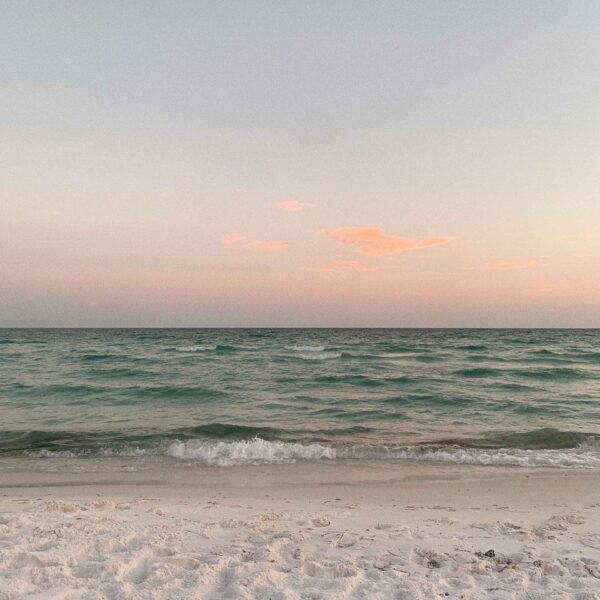 This image shows a tranquil beach scene at twilight with gentle waves approaching the shore and pastel hues in the sky lightly touched by clouds.