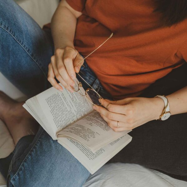 A person sits comfortably, holding glasses and reading a book. They wear a casual orange shirt, blue jeans, and have a wristwatch on.