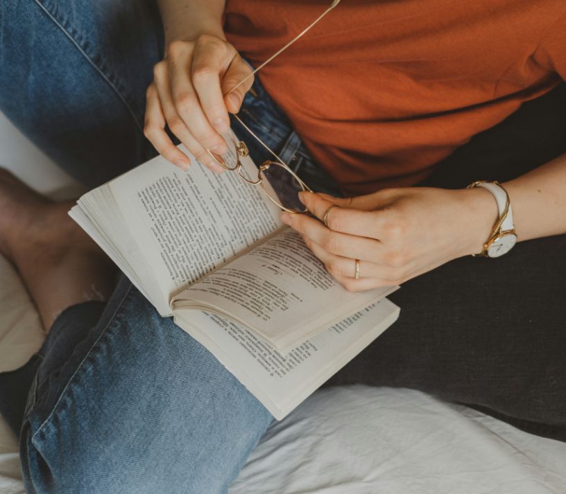 A person is seated, holding a pair of glasses and an open book. They wear a red shirt, blue jeans, and a wristwatch, suggesting a moment of relaxation.