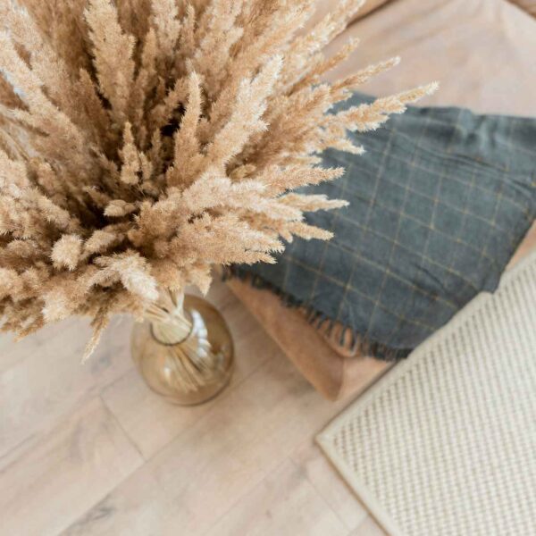 A cozy interior scene with a vase of pampas grass on a wooden floor beside a textured cushion and a dark patterned throw pillow.