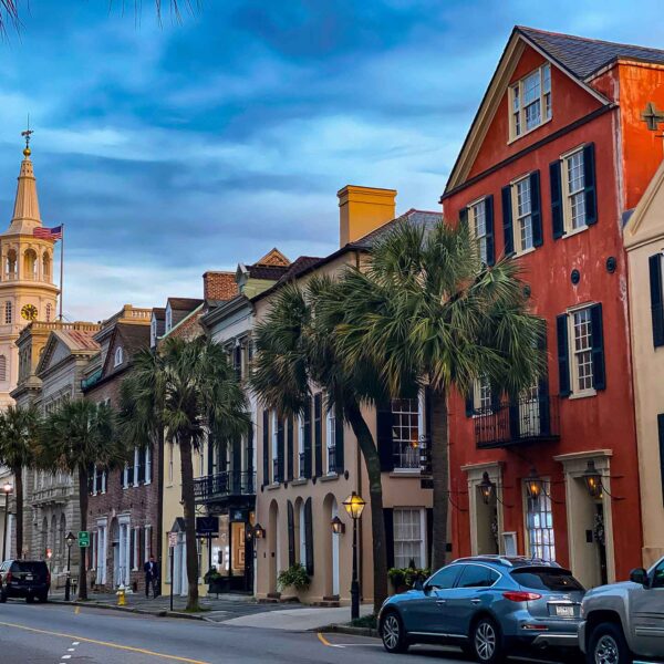 Historic street lined with colorful, traditional buildings and palmetto trees under a dusk sky, with a steepled church tower rising in the background. Cars parked curbside.