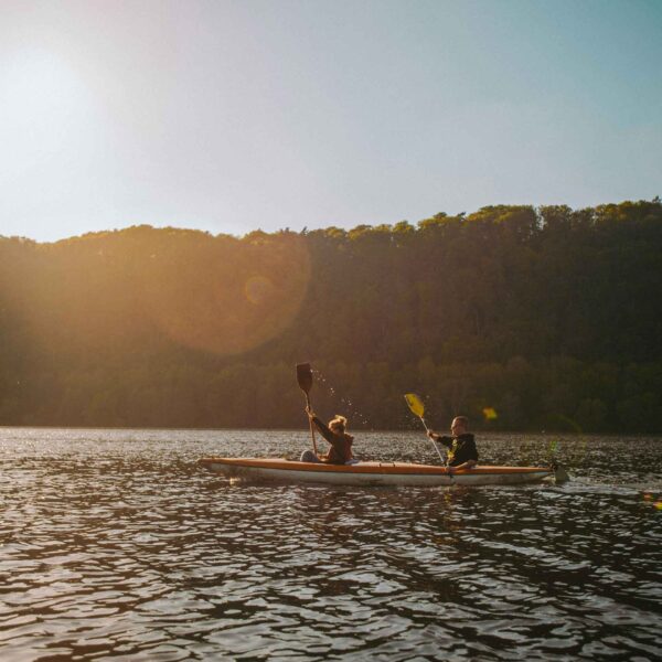 Two people are kayaking on calm water during sunset. Trees line the shore behind them, and the sun creates a warm, tranquil atmosphere.