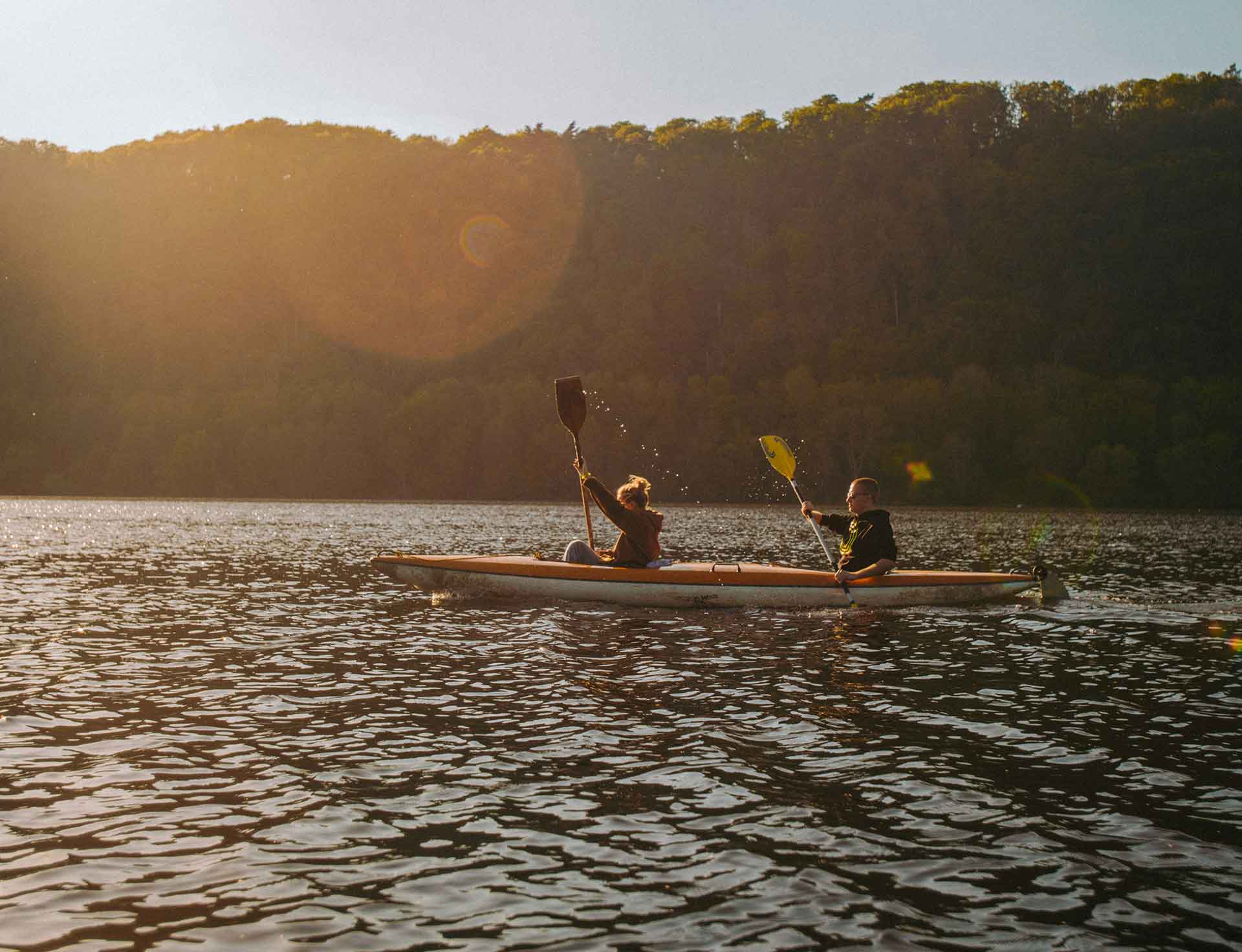 Two people are kayaking on a calm water body against a backdrop of forested hills during sunset, which casts a warm glowing light and lens flare.