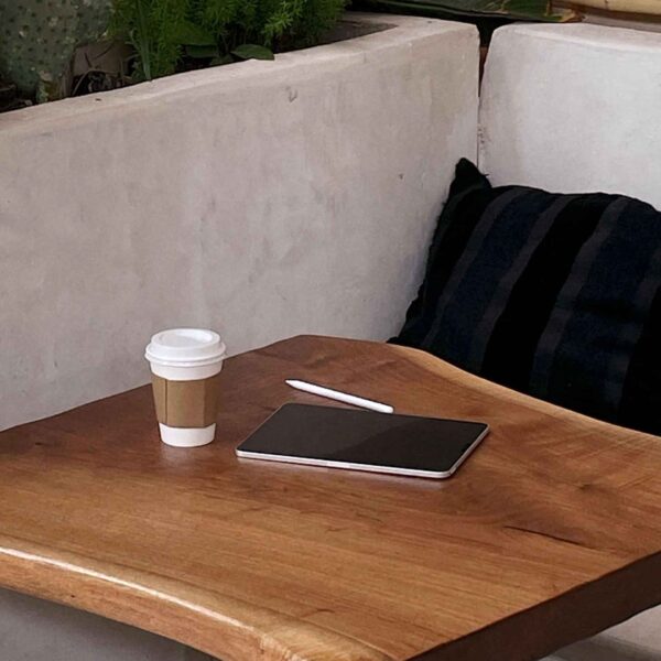 A cozy corner with a wooden table, a disposable coffee cup, a closed laptop, a cushion, and plants in the background, suggesting a relaxed atmosphere.