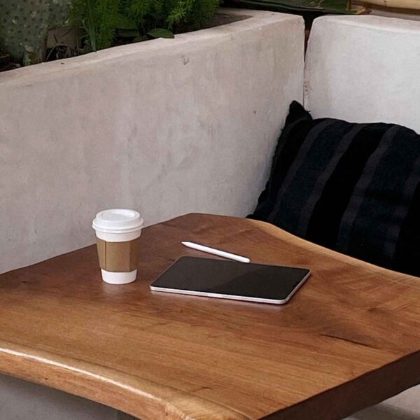 A cozy café corner with a wooden table, featuring a disposable coffee cup and a smartphone. Lush plants and a cushioned bench enhance the tranquil ambiance.