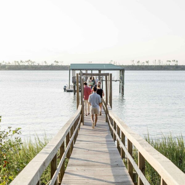 Three people walk towards a covered dock extending over calm water, with lush foliage to the sides, under a clear sky.