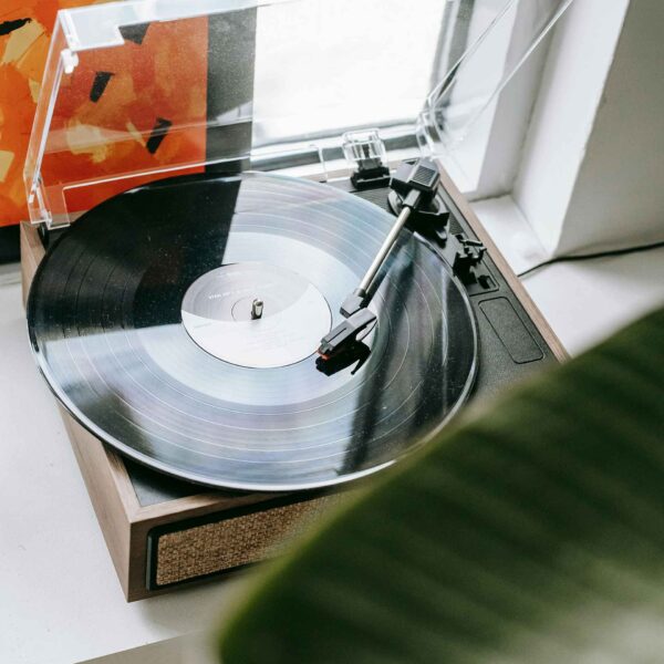 A turntable by a window plays a vinyl record, with an album cover visible, and a plant's leaf in the foreground partially obscuring the view.