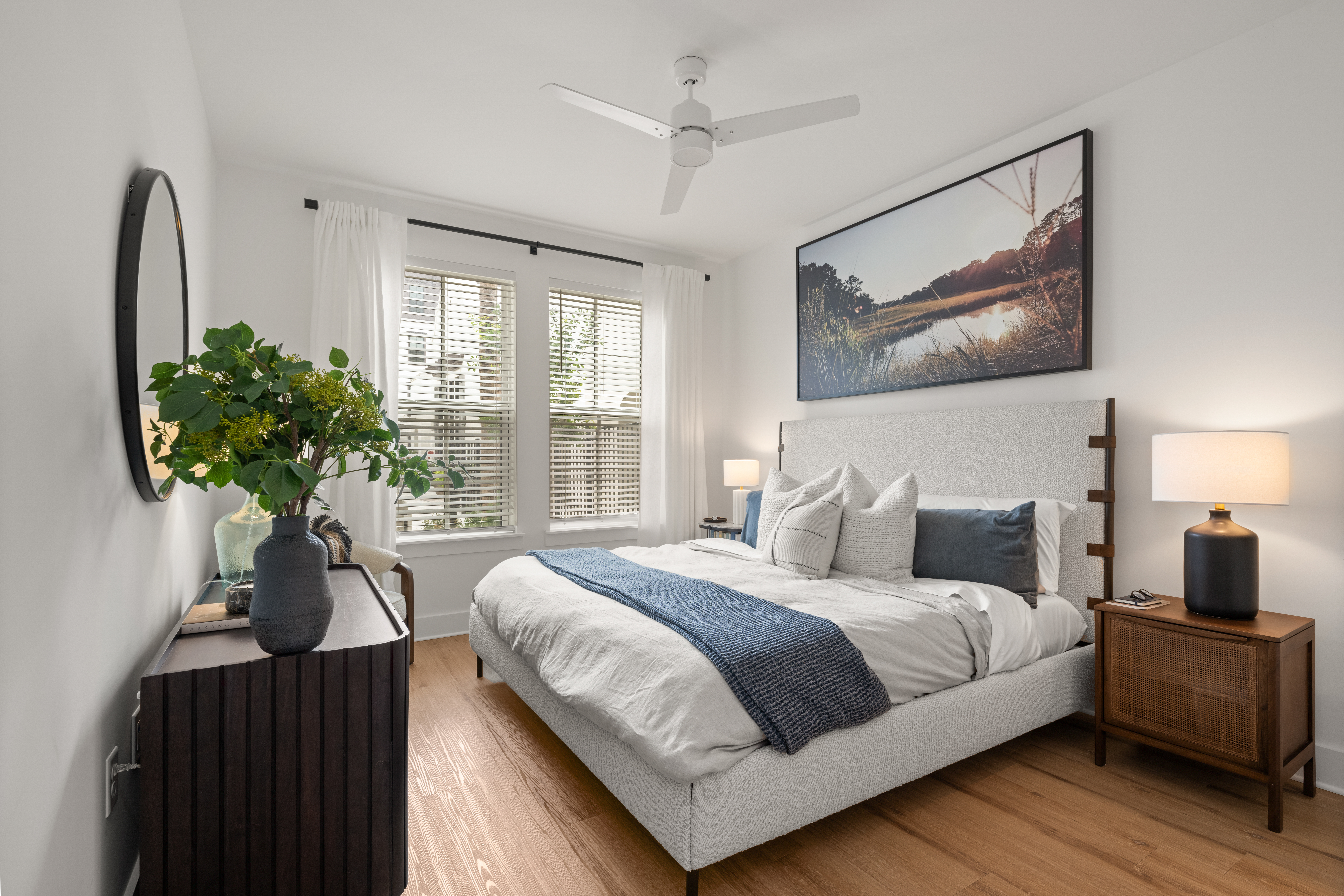 Bright bedroom featuring white bedding, two bedside tables with lamps, and a window with white curtains.
