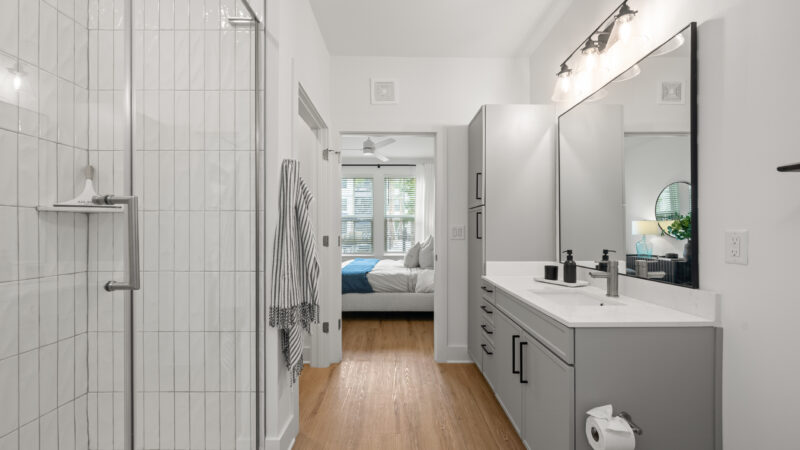 Bright bathroom featuring white tile shower walls, gray cabinetry, and a wide mirror above the sink.